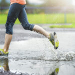 young-woman-running-on-asphalt-sports-field-in-rainy-weather-details-of-legs-and-sport-SBI-305121763