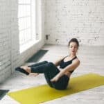 exhausted-young-european-woman-performing-boat-asana-while-practicing-yoga-indoors-brunette-girl-sitting-green-mat-doing-navasana-pose-with-back-legs-lifted-arms-extended-forward