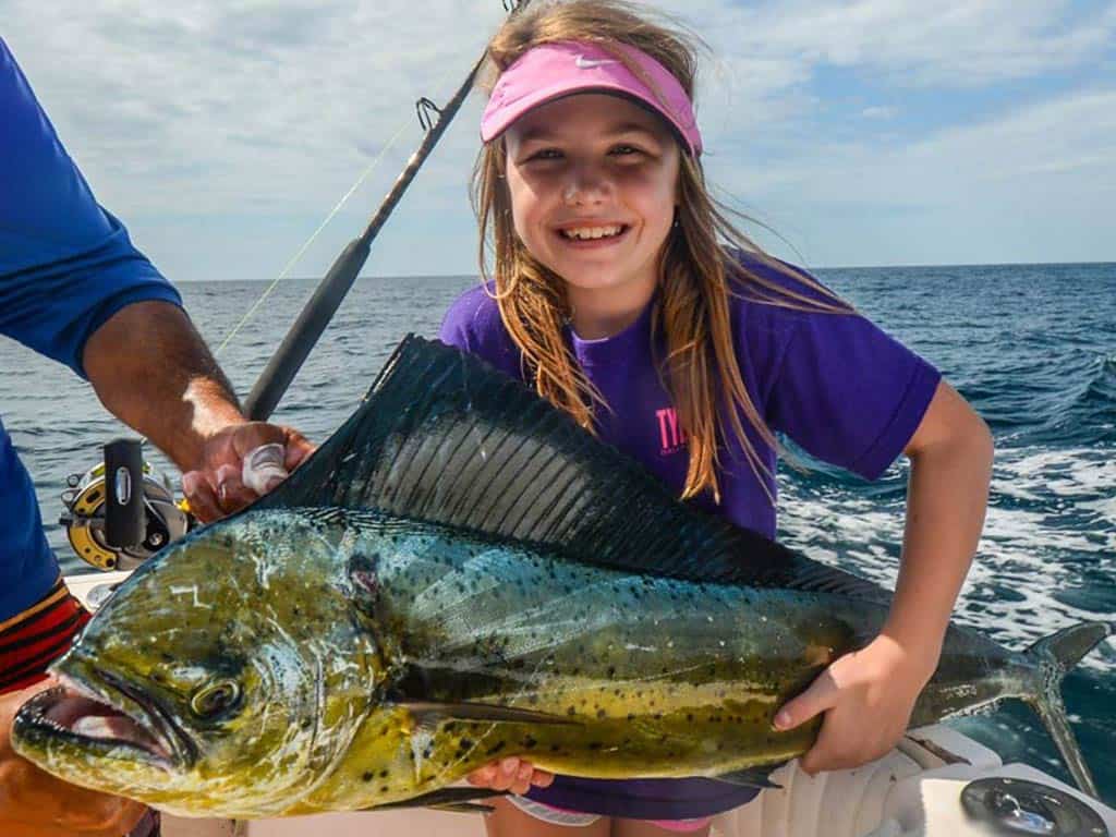 A photo of a little girl smiling and holding Mahi Mahi with both hands while standing on a Coco charter boat